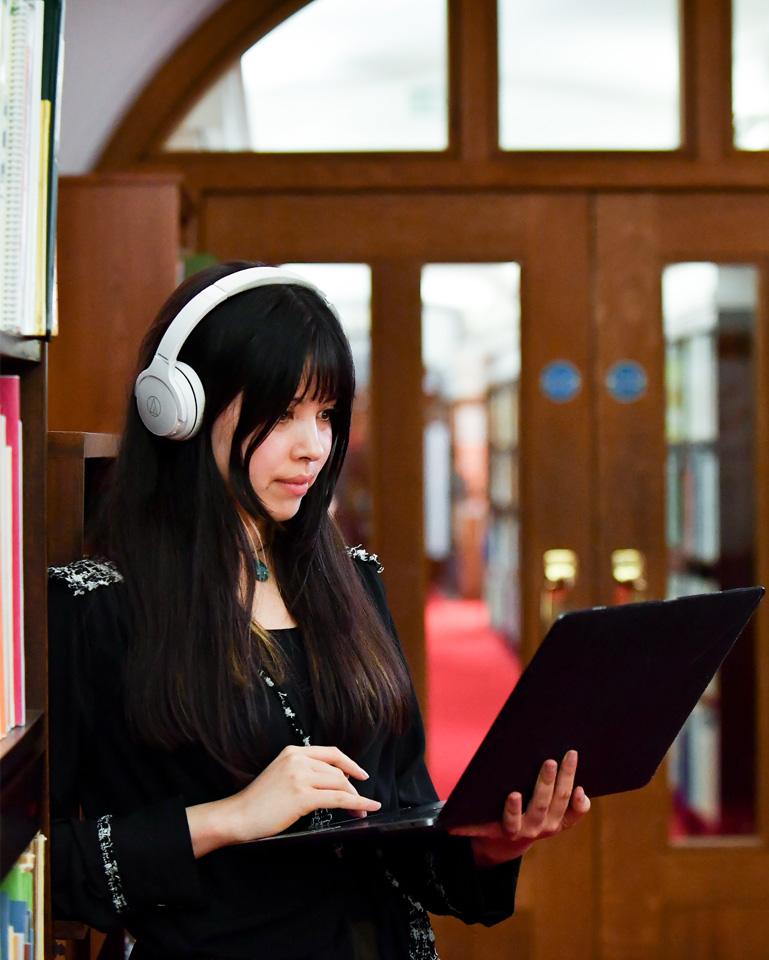 A student with long, dark hair, wearing headphones, looking on a laptop.